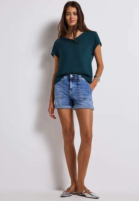 Frau mit grünem Shirt und Jeansshorts steht vor weißer Wand.